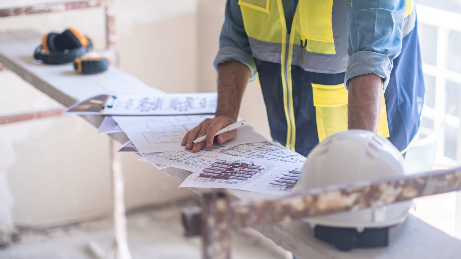 Worker in vest editing and correcting renovation project papers stands near construction equipment and protective hardhat man in overall working in unfinished living room
